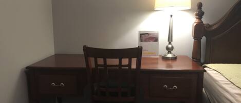 Una habitación pequeña y tenuemente iluminada, con un escritorio de madera, una silla, una lámpara y una cama con una manta verde.