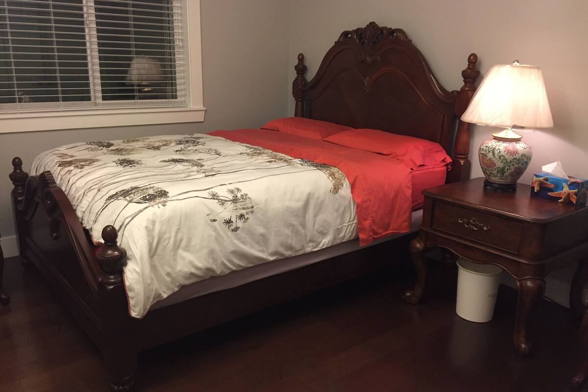 A neatly made bed with a floral bedspread and red pillows, a wooden nightstand with a lamp and a tissue box, and a window with blinds.