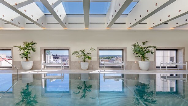An indoor swimming pool with clear glass walls, potted plants, and a view of the cityscape. - Barceló Torre de Madrid (Madrid)