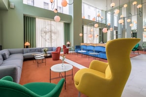 Un área de descanso moderna con un sofá modular, un sillón amarillo y un sofá azul. Hay varias mesas y sillas, y la habitación tiene ventanas amplias con cortinas.