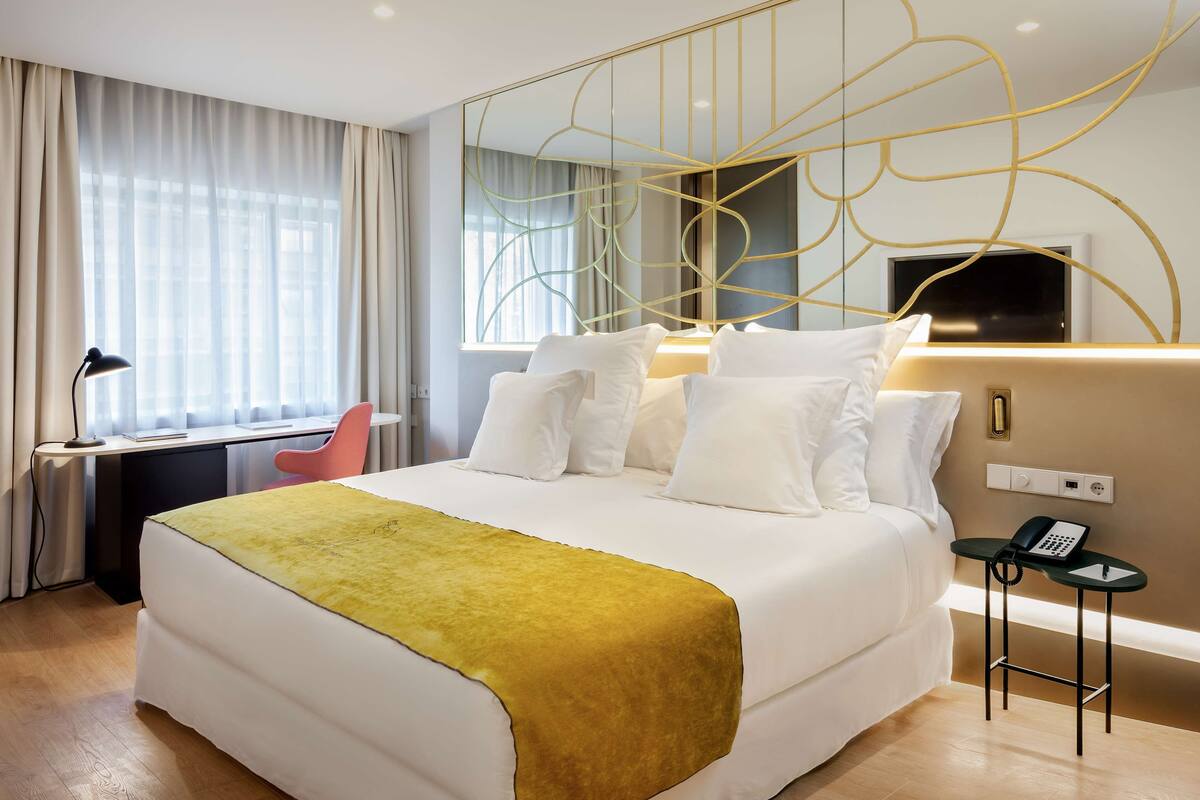 Habitación de hotel con cama, escritorio, silla, mesita de noche con teléfono y un espejo grande con diseño geométrico.