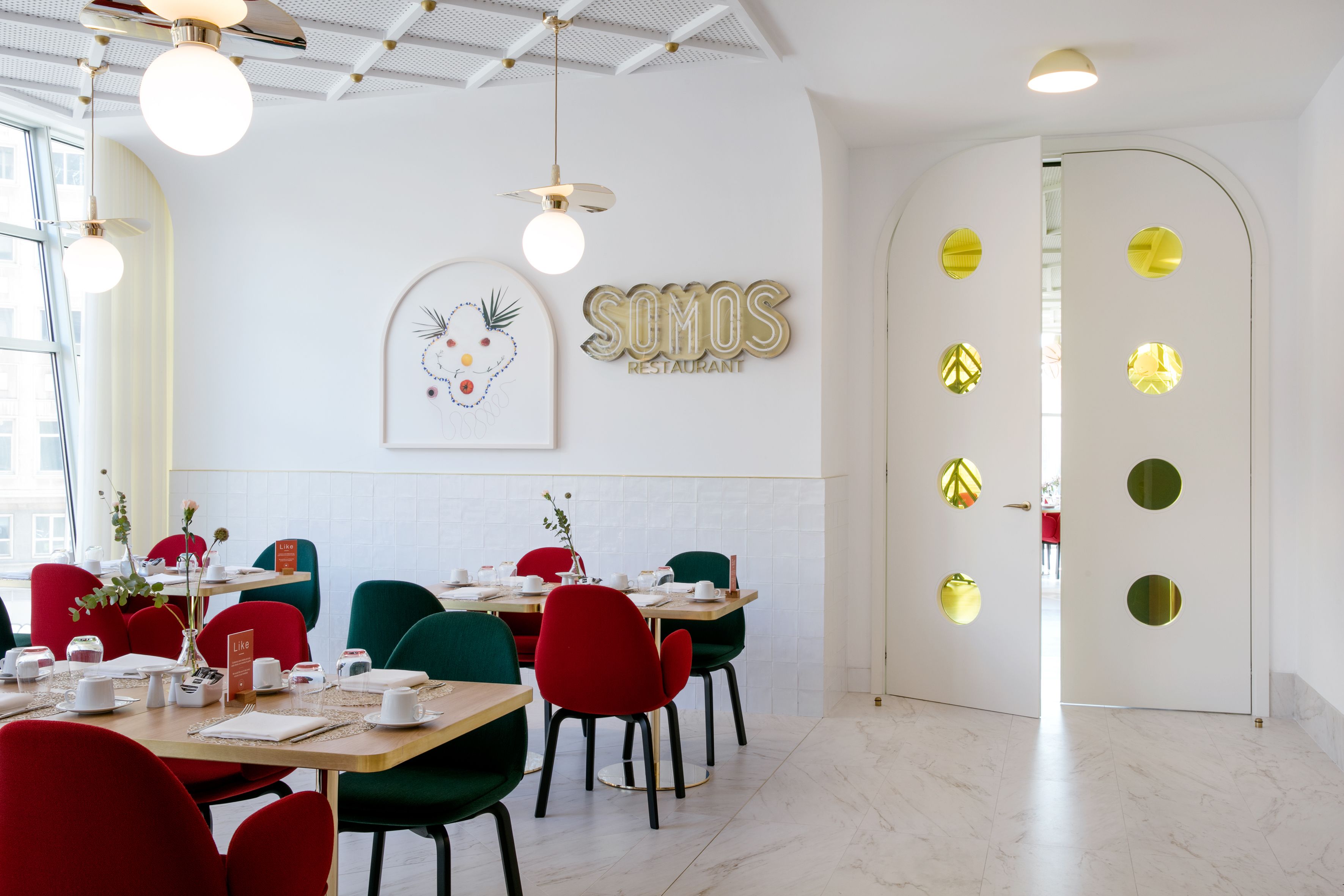 A modern restaurant interior with round tables, red and green chairs, and a white wall with a 'SOMOS RESTAURANT' sign.
