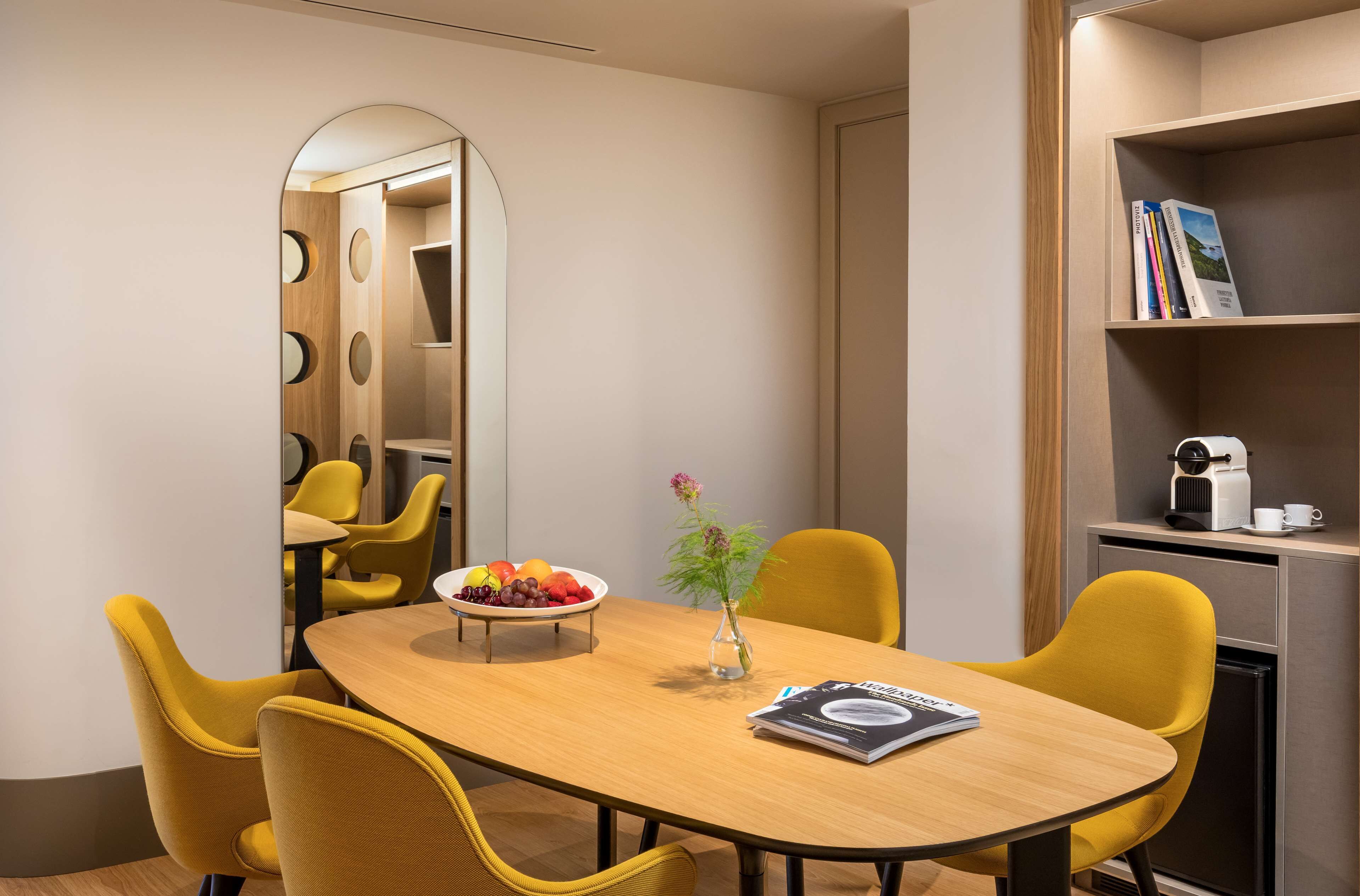 Una sala de conferencias moderna con una mesa de madera, sillas amarillas, un frutero, un florero y una estantería con libros y una máquina de café.