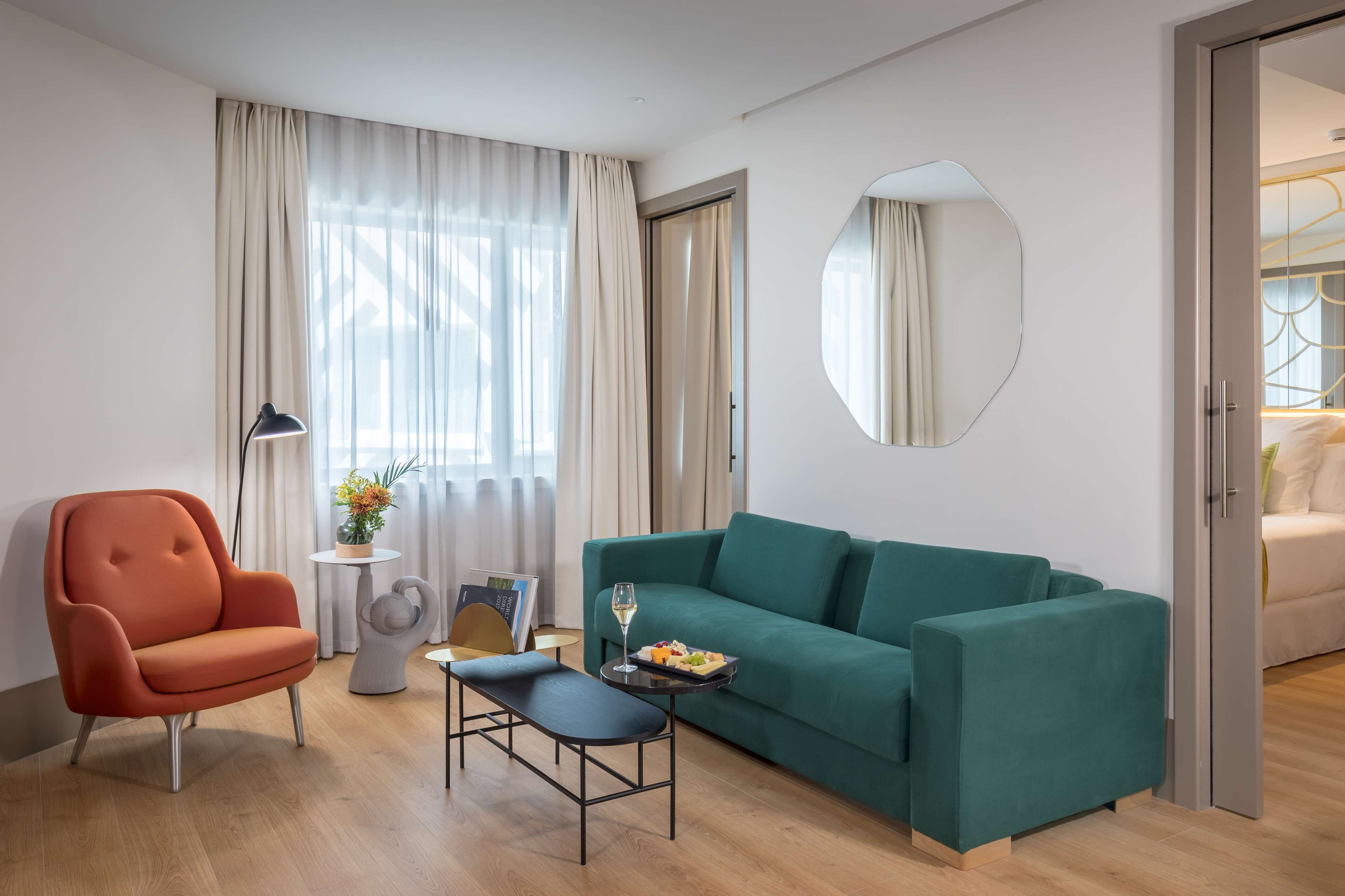 Habitación de hotel moderna con sofá color turquesa, piso de madera, espejo redondo y mesa de comedor con una copa de vino y fruta.