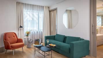 Habitación de hotel moderna con sofá color turquesa, piso de madera, espejo redondo y mesa de comedor con una copa de vino y fruta.