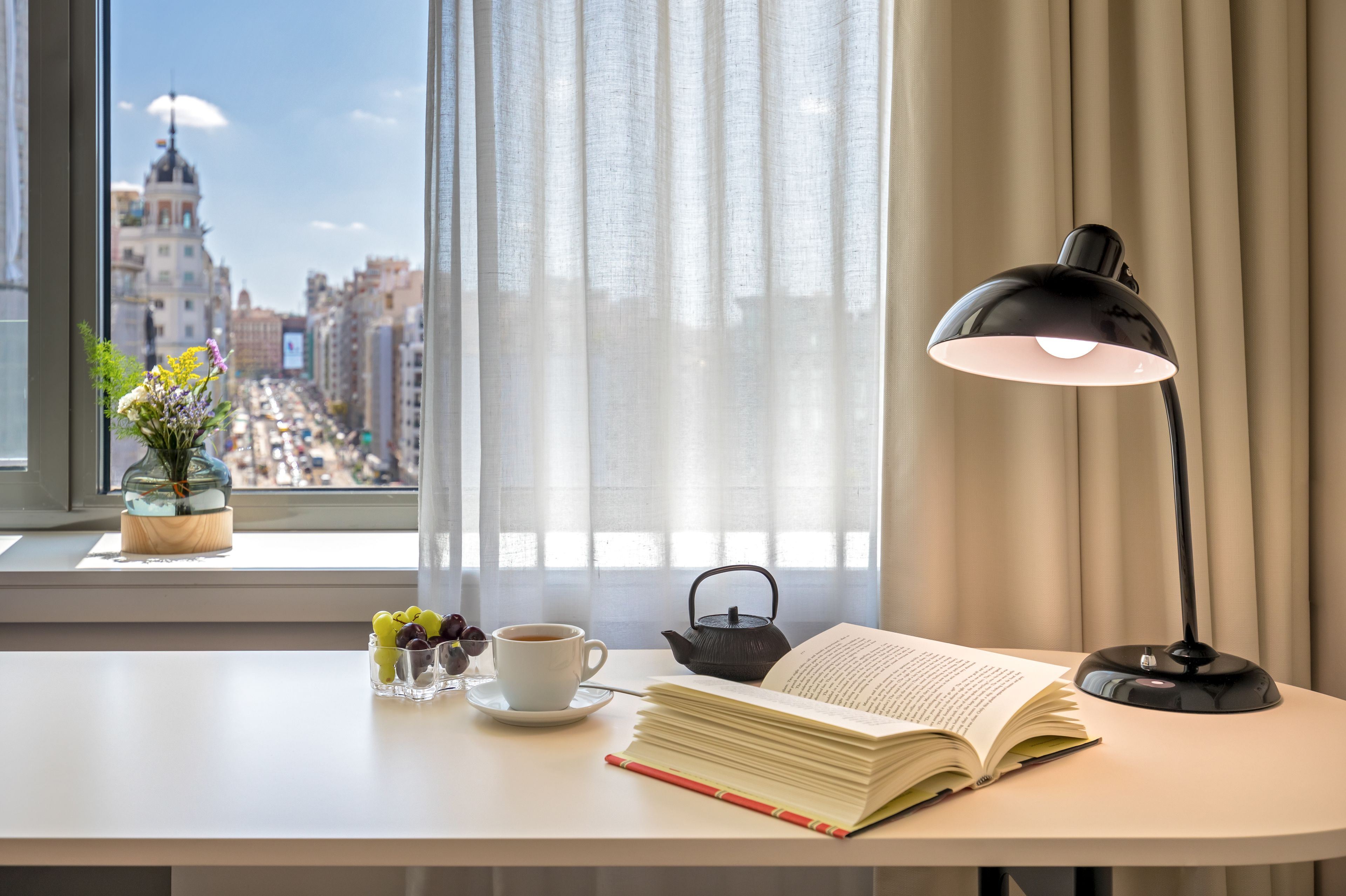 A neatly arranged desk with a book, a cup, a teapot, and a vase of flowers by a window with a city view.