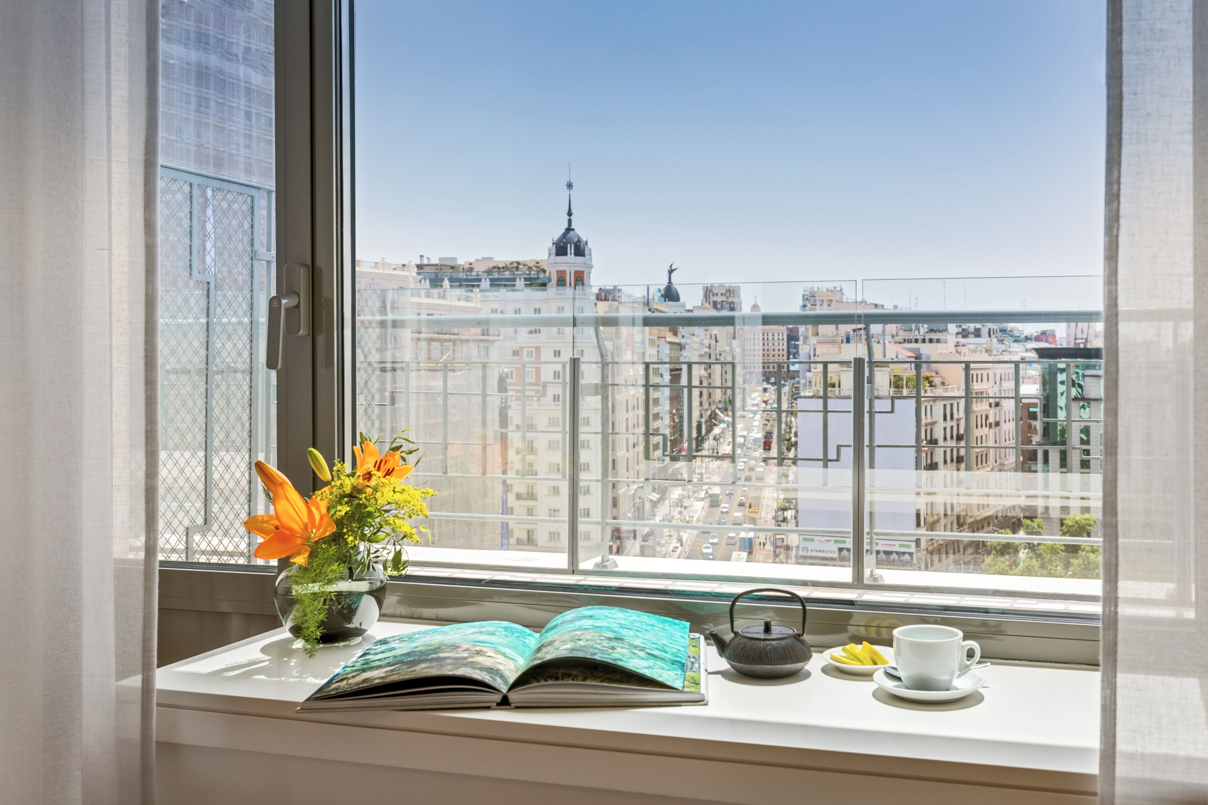 A window with a view of a cityscape, a potted plant with flowers, an open book, a teapot, and a cup on a windowsill.
