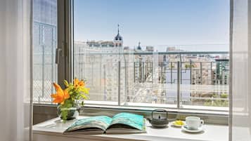 A window with a view of a cityscape, a potted plant with flowers, an open book, a teapot, and a cup on a windowsill.