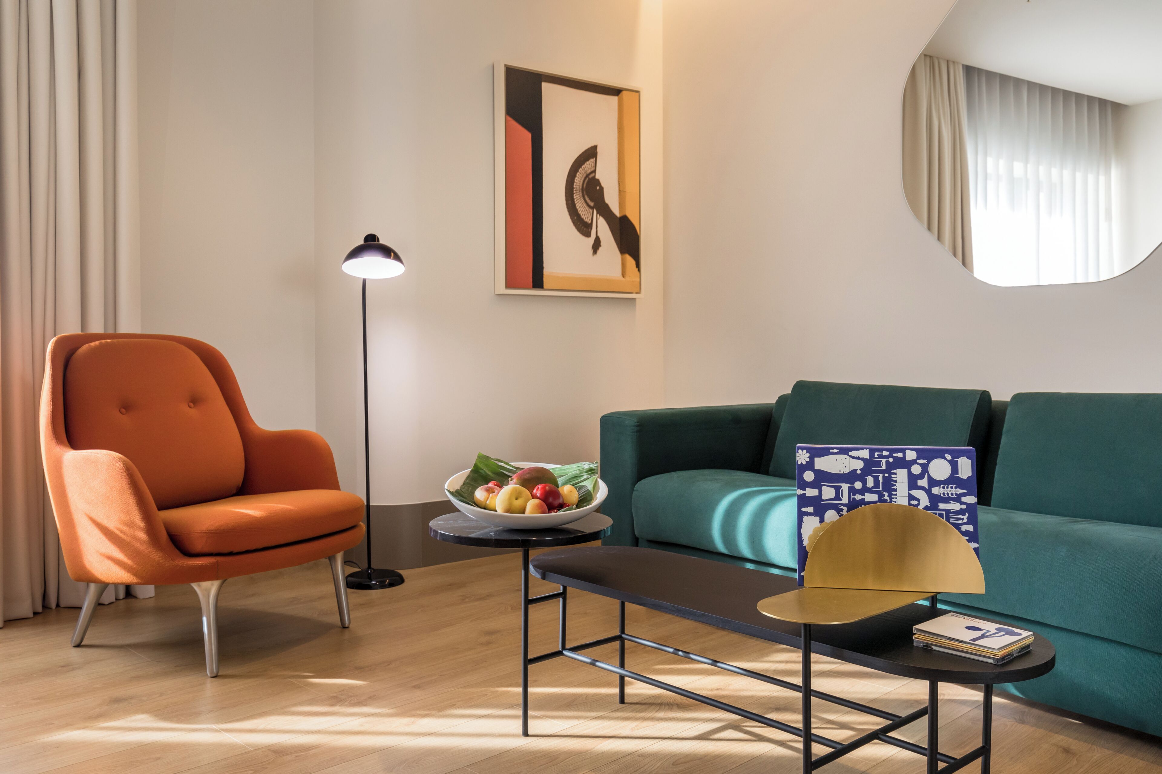Una sala de estar moderna con un sofá color turquesa, un sillón naranja, una lámpara de pie, una mesita con un frutero y una obra de arte enmarcada en la pared.
