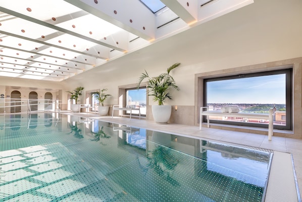 Una piscina interior moderna con paredes de vidrio transparente, ventanas amplias y plantas en macetas.