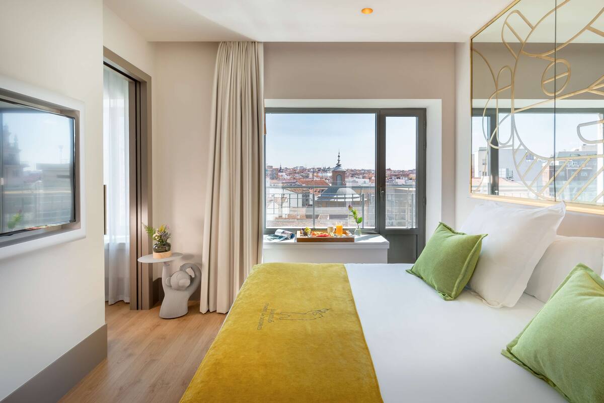 Habitación de hotel con una cama grande, vistas a la ciudad, una mesita con un jarrón y una ventana con cortinas.