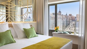 A hotel room with a large bed, green pillows, a green throw blanket, a view of a cityscape through large windows, and a decorative mirror with geometric patterns. - Barceló Torre de Madrid (Madrid)
