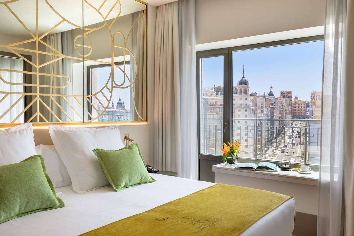 Habitación de hotel con una cama grande, almohadas verdes, un plaid verde, vistas de un paisaje urbano a través de ventanas amplias y un espejo decorativo con patrones geométricos.