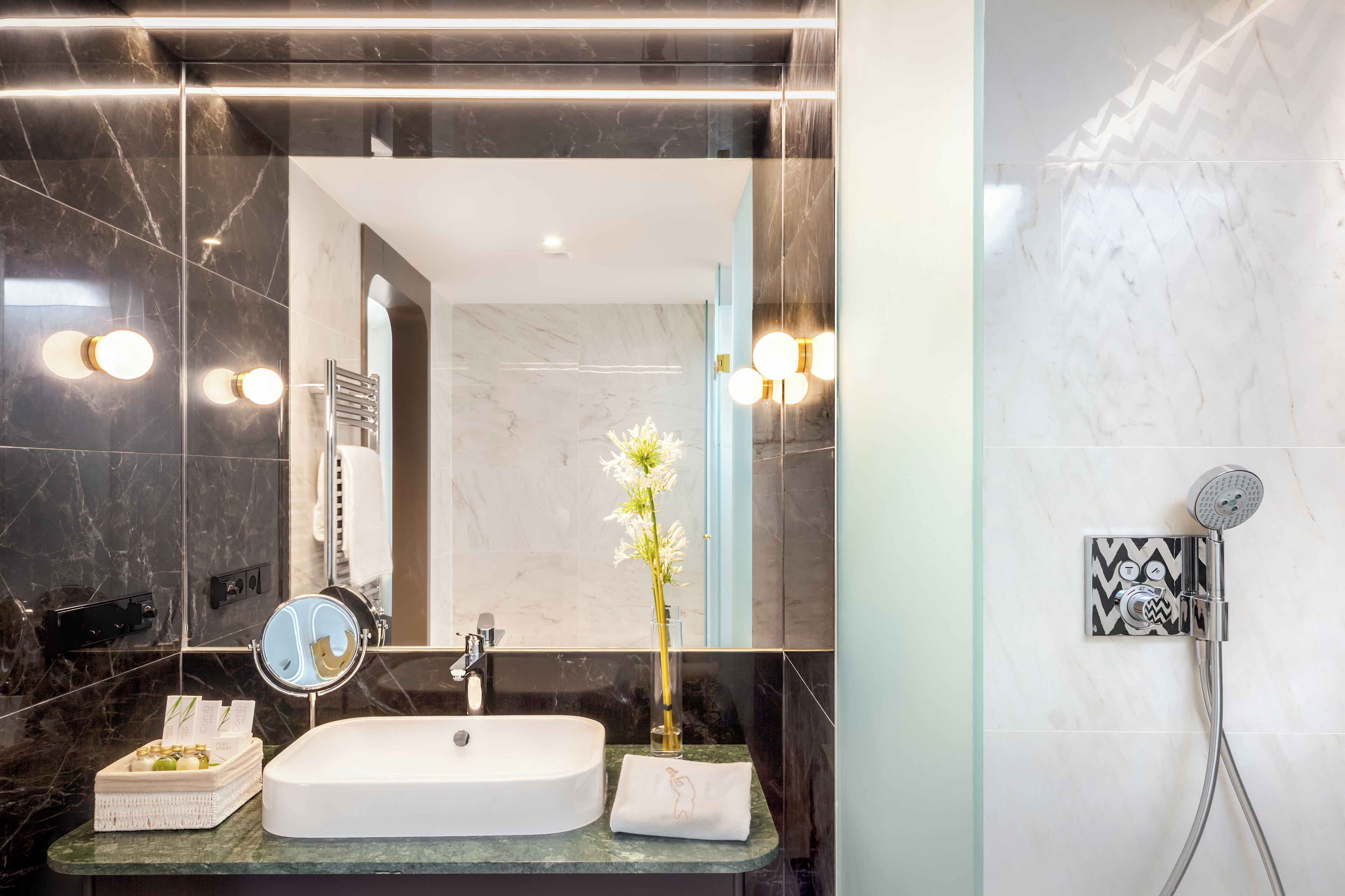 Une salle de bain avec un lavabo blanc, un miroir, une serviette, un vase de fleurs et une douche.
