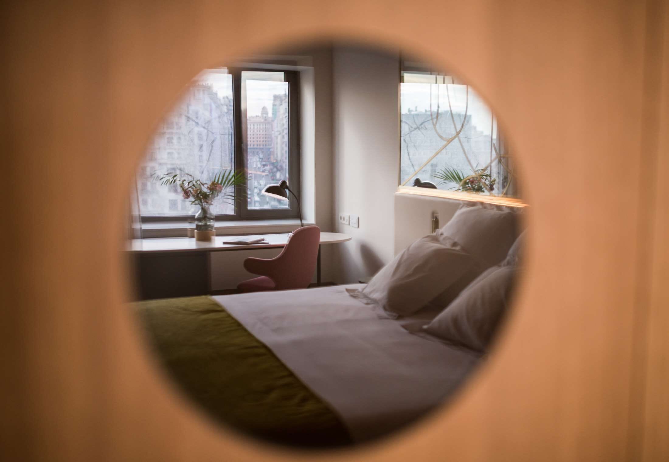 A bedroom with a bed, a desk, a chair, and a window with a view of buildings.
