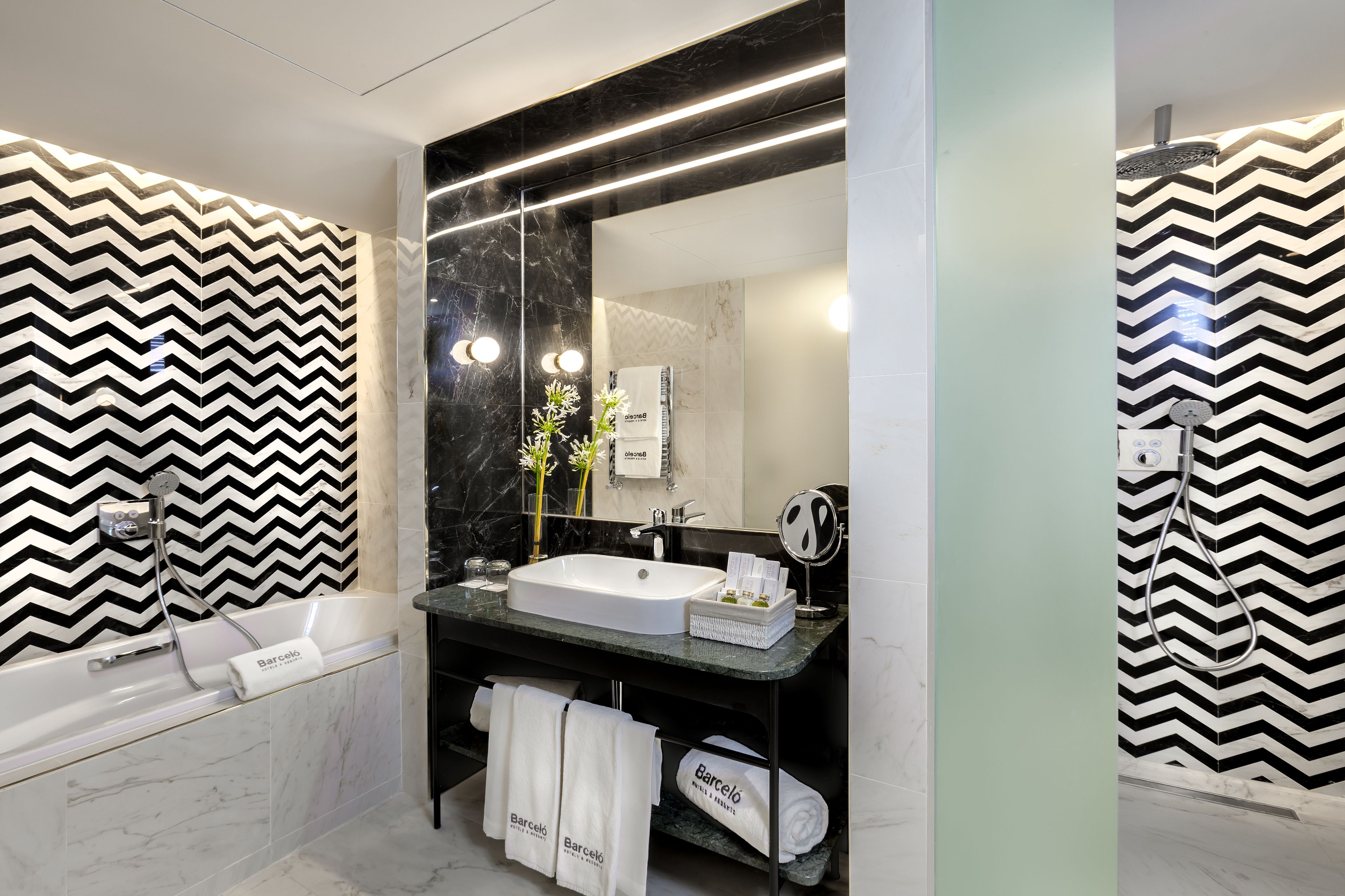 Un baño moderno con una pared de diseño en blanco y negro con patrón de espiga, un lavabo blanco, un espejo grande, un cabezal de ducha de mano y una bañera.