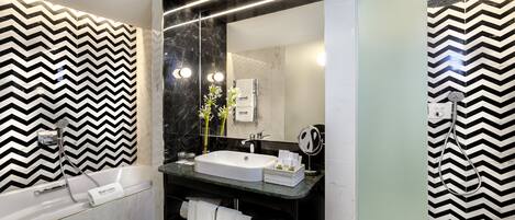 Un baño moderno con una pared de diseño en blanco y negro con patrón de espiga, un lavabo blanco, un espejo grande, un cabezal de ducha de mano y una bañera.