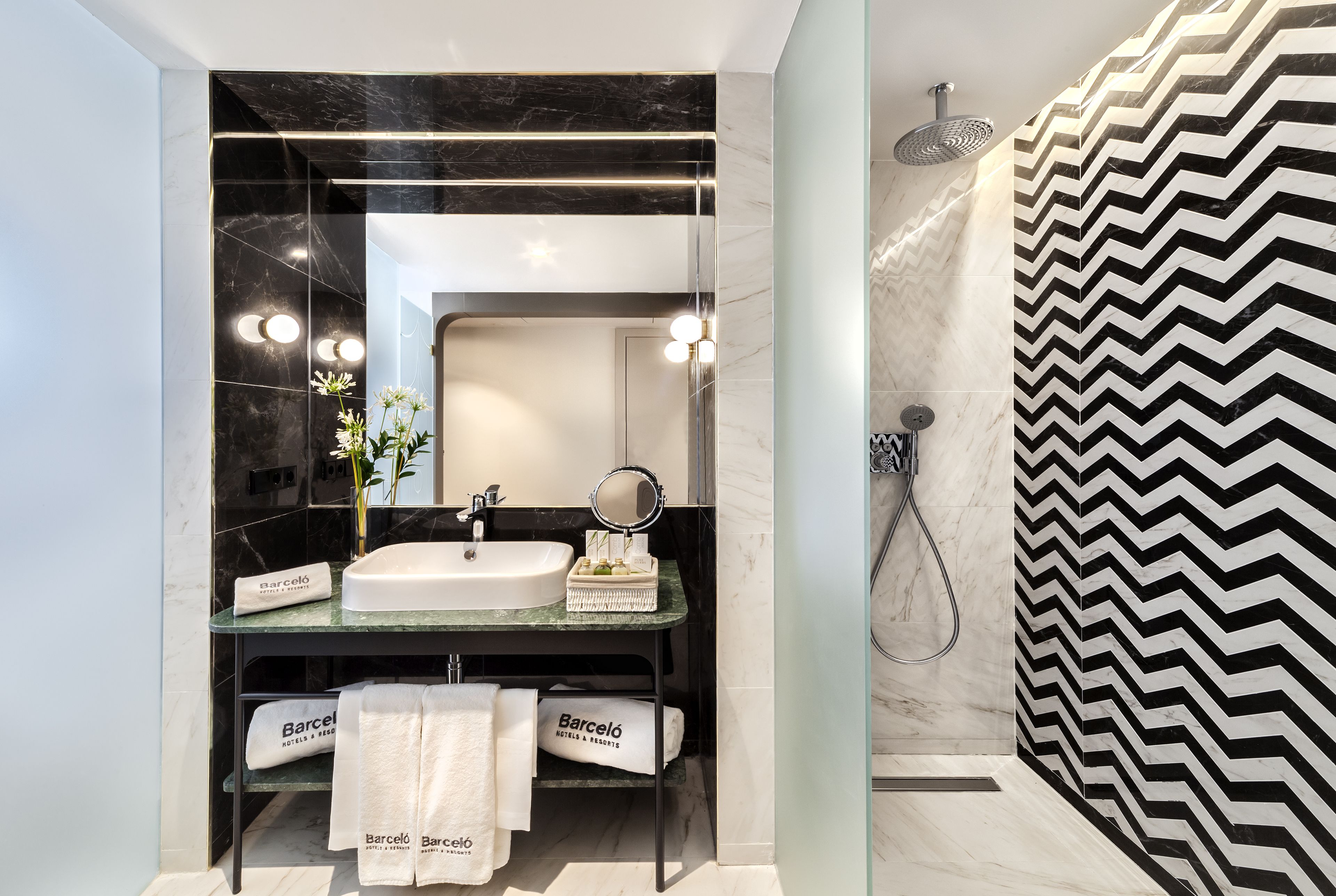 Un baño moderno con un espejo grande, un lavabo y una ducha. Hay toallas y una pequeña cesta en la encimera.