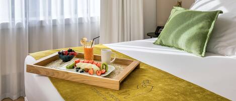 A hotel room with a bed, a tray with breakfast including fruits and juice, a green pillow, and a wall-mounted nightstand.