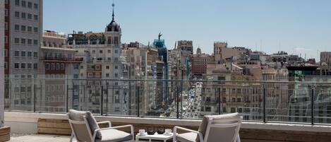 A rooftop terrace with white chairs and a table, overlooking a cityscape with various buildings and a prominent blue structure.