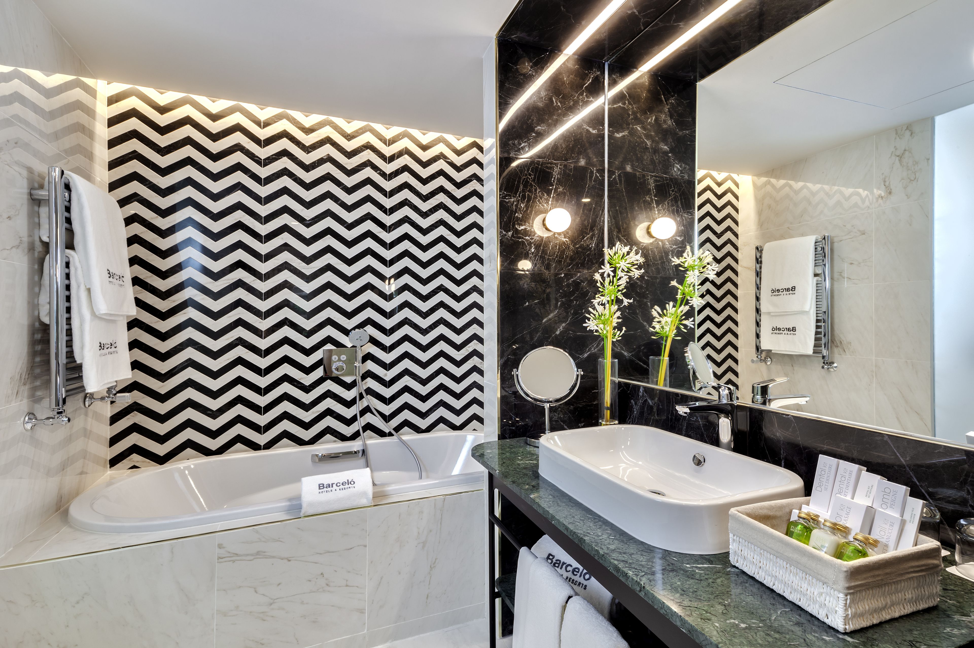 A modern bathroom with a zigzag patterned wall, a large mirror, a bathtub, and a sink with toiletries.
