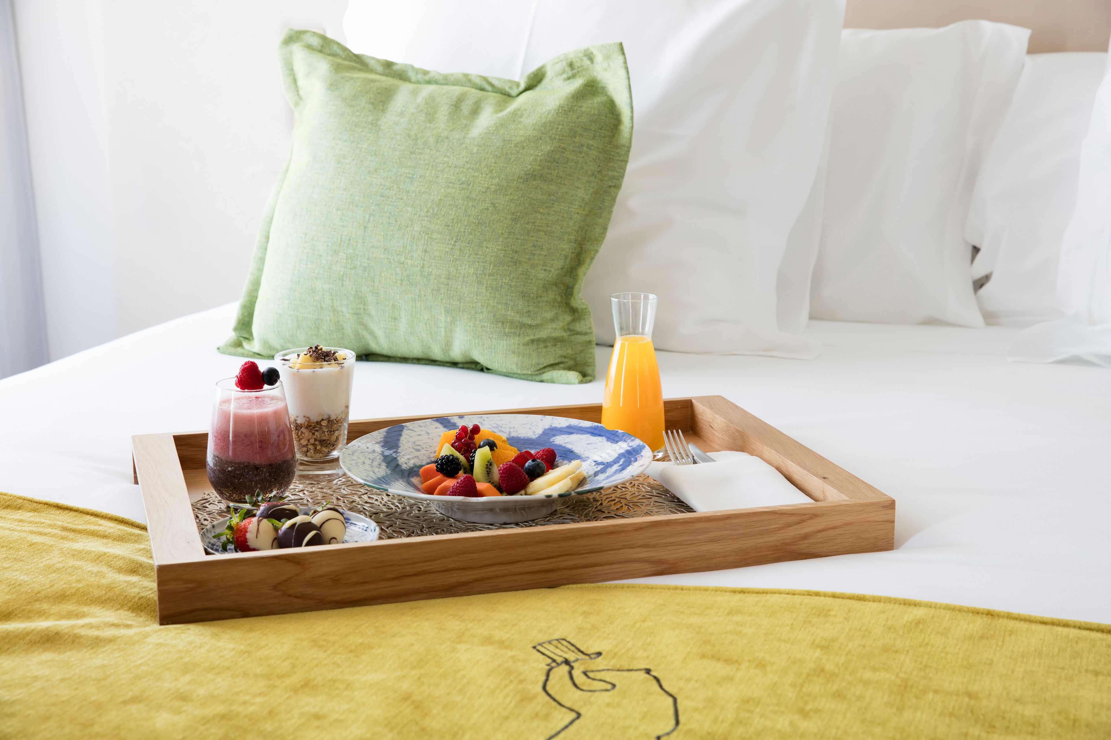 Bandeja de desayuno con fruta, yogur y jugo sobre una cama con ropa blanca y almohada verde.