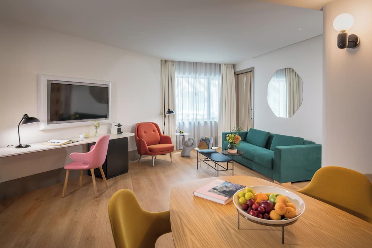 Una sala de estar moderna con un sofá color turquesa, un sillón rosa, una mesa de comedor de madera con un bol de fruta, un escritorio con una computadora y un espejo grande en la pared.