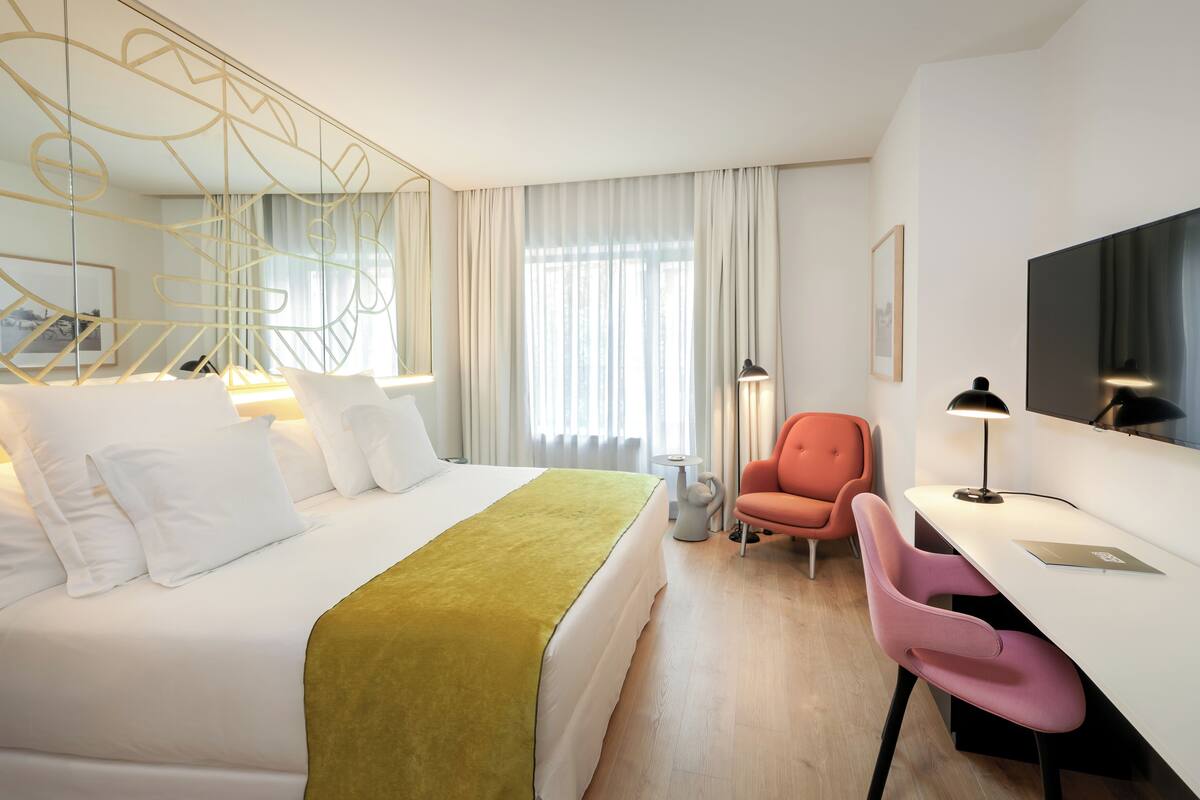 Habitación de hotel con cama, escritorio, silla, televisor y ventana con cortinas.