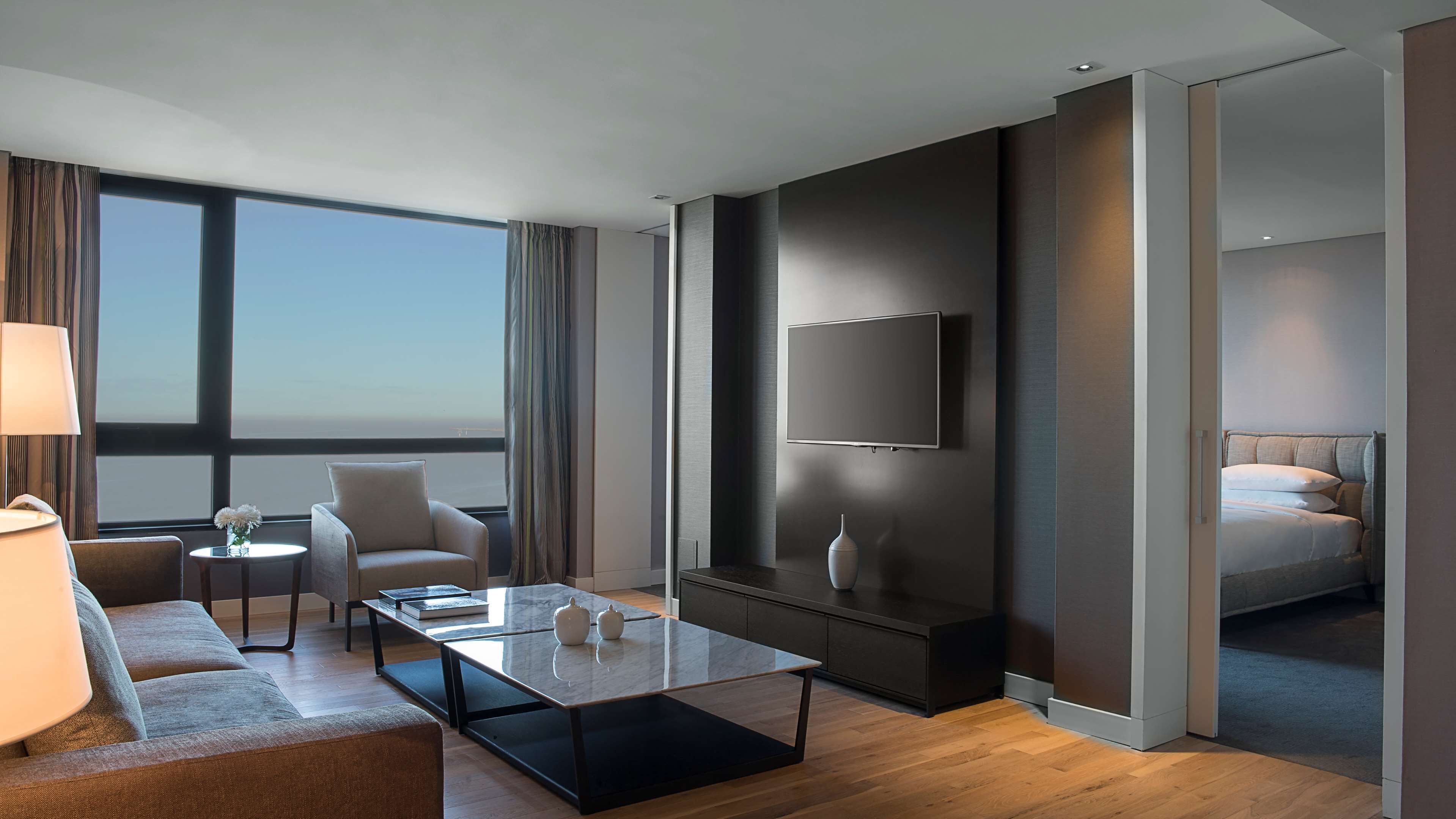 Una habitación de hotel moderna con un ventanal grande, un televisor de pantalla plana, un sofá, una mesa de centro y una cama.