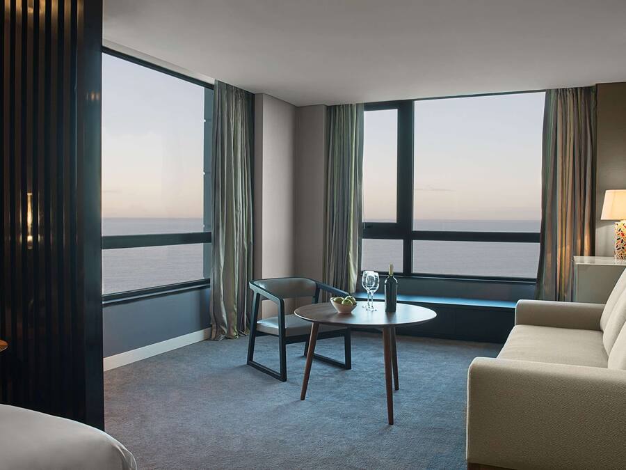 Habitación de hotel con un gran ventanal que ofrece vistas al mar, una mesa redonda con una silla, un sofá y una lámpara sobre una mesita auxiliar.