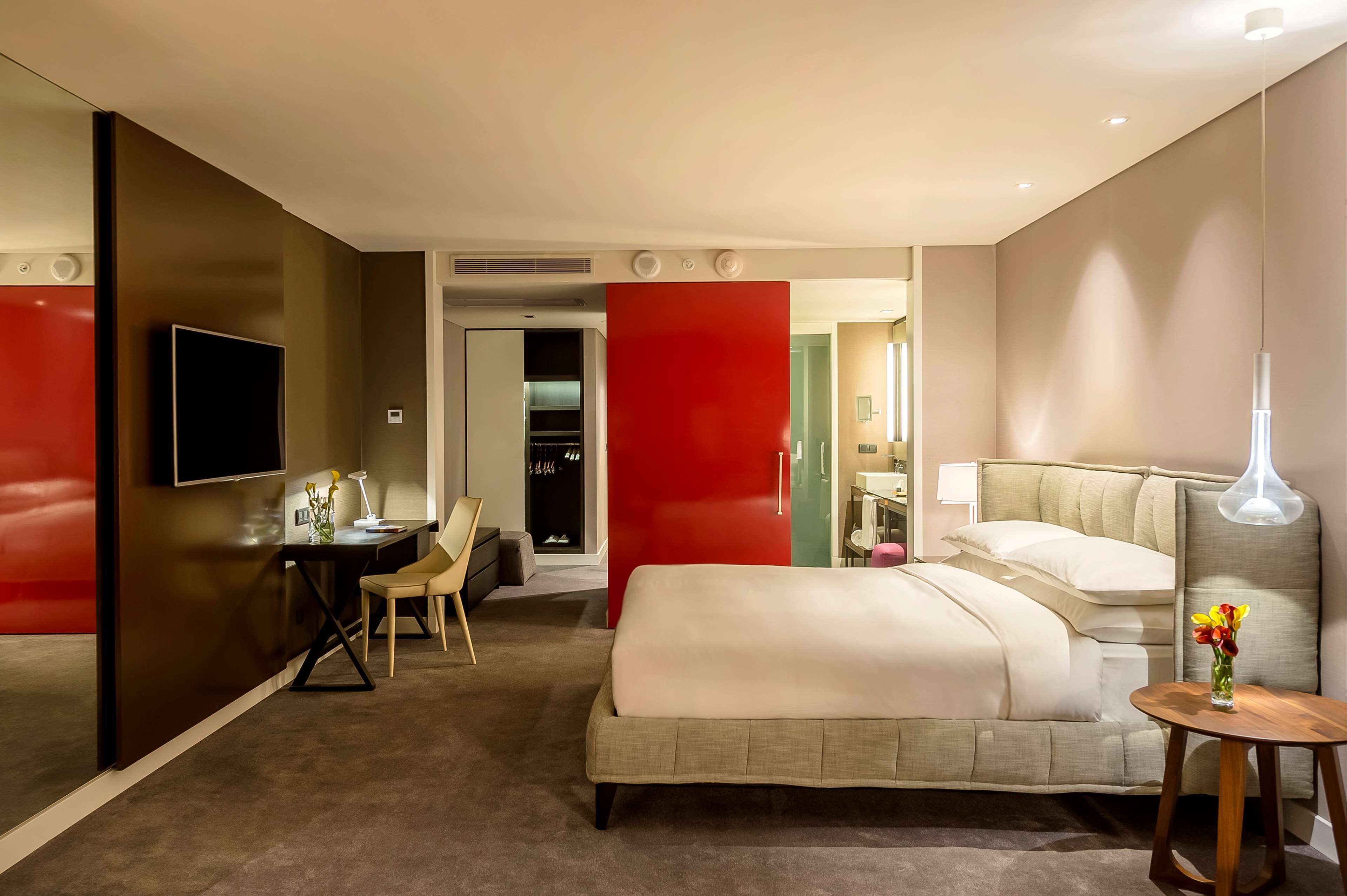 A modern hotel room with a large bed, a desk with a chair, a TV, and a red accent wall.
