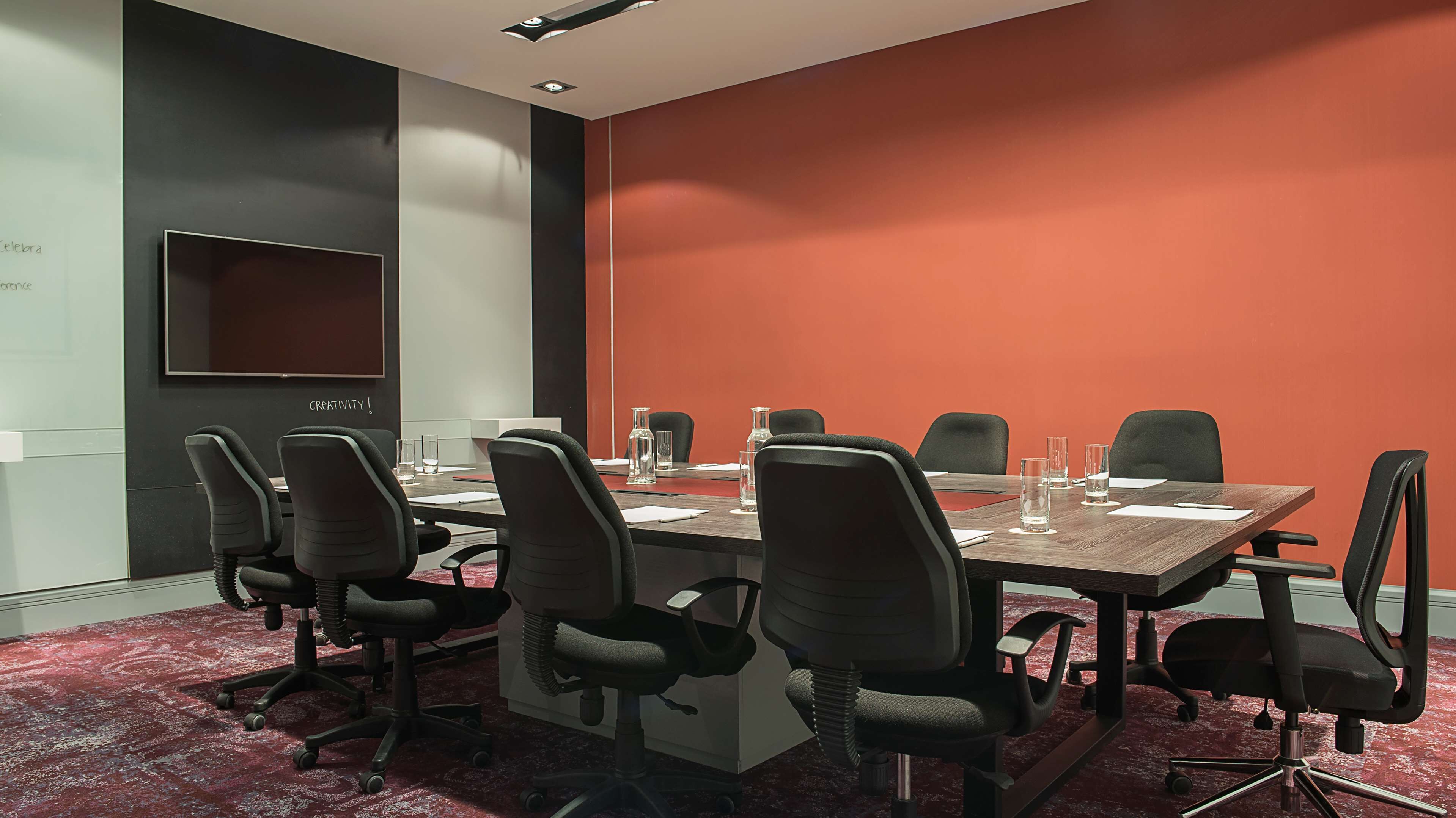 A conference room with a long table, six chairs, and a large screen on the wall.