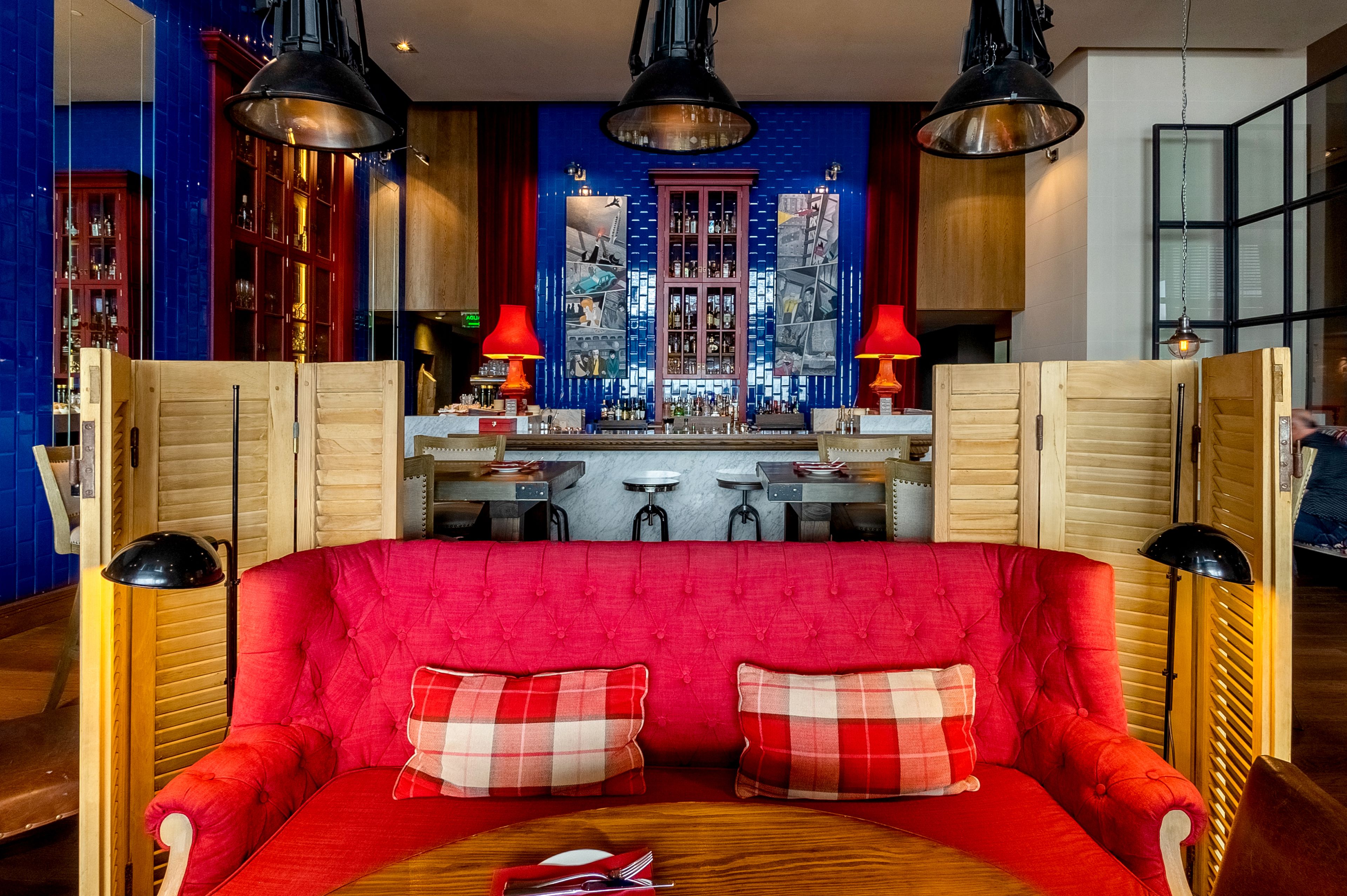Un sofá rojo con almohadones a cuadros, una mesa de madera y una zona de bar con taburetes y lámparas rojas.
