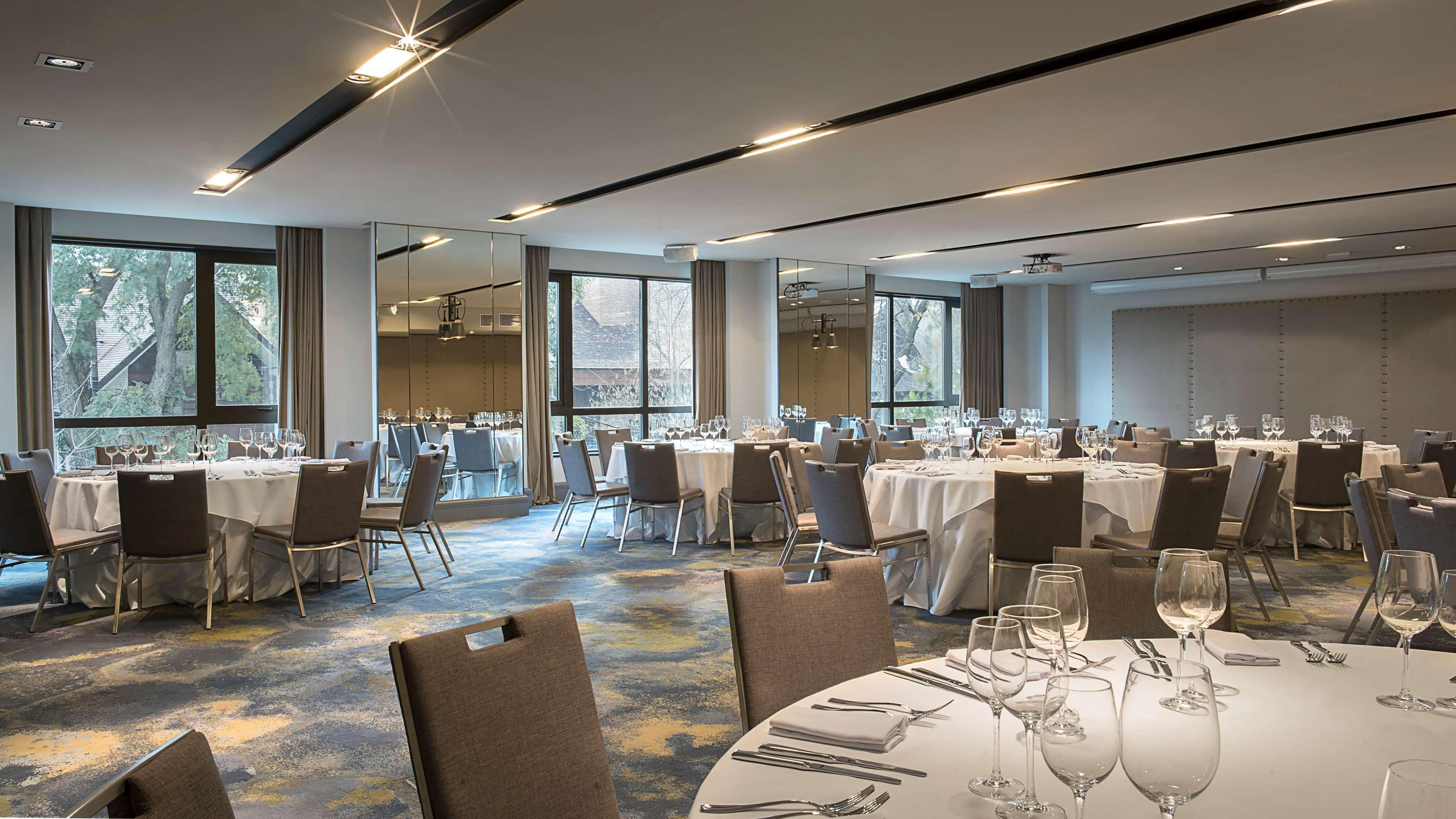 A spacious banquet hall with multiple round tables set for dining, large windows offering natural light, and a modern ceiling design.