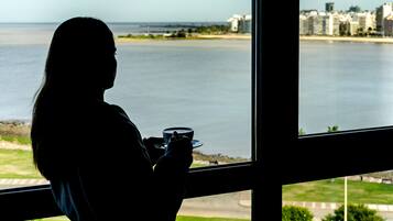 Silueta de una persona sosteniendo una taza, vista a través de una ventana con vista a un cuerpo de agua y un horizonte urbano lejano.