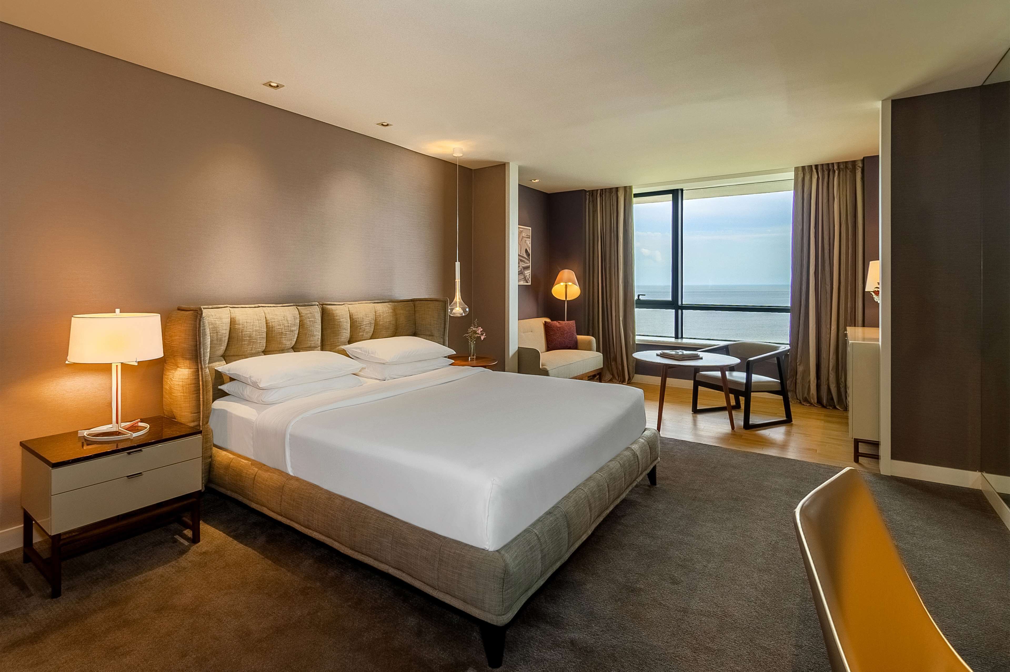 A hotel room with a large bed, a nightstand, a desk, a chair, and a view of the sea.