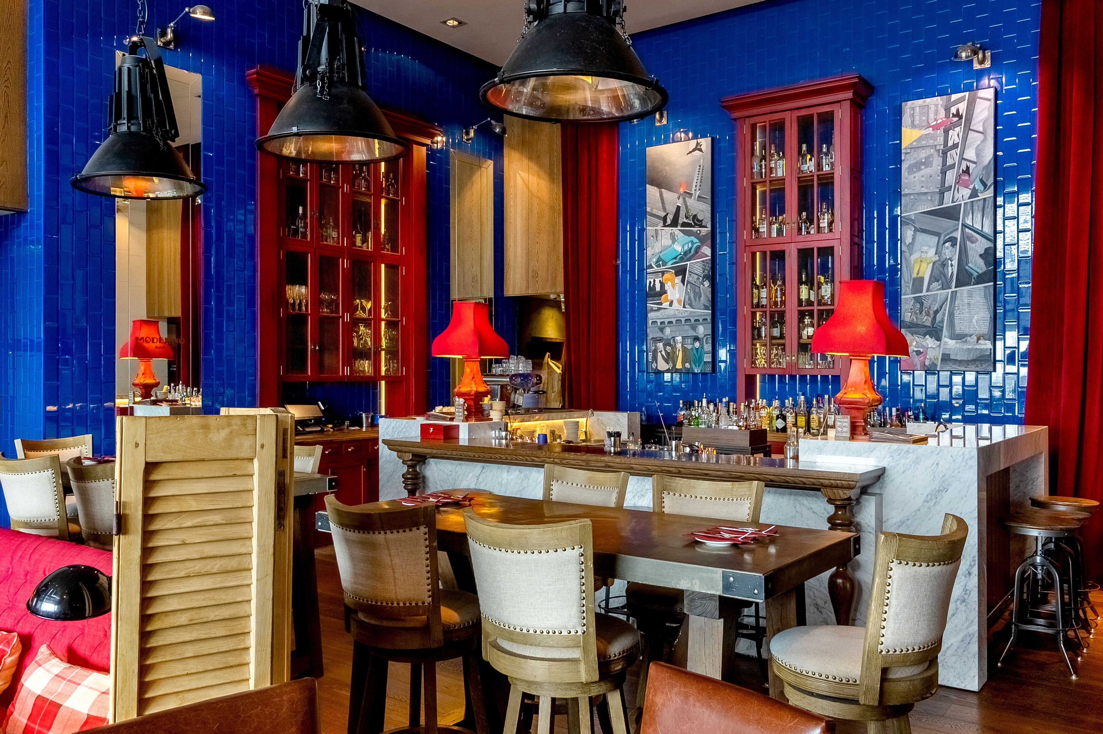 Zona de bar con mobiliario de madera, lámparas colgantes rojas y negras, y una barra con una variedad de botellas.