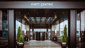 The entrance of the Hyatt Centric hotel with a modern design, featuring a large glass facade, a central entrance with revolving doors, and potted plants.