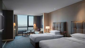 A hotel room with two beds, a desk, a chair, and a view of the cityscape.