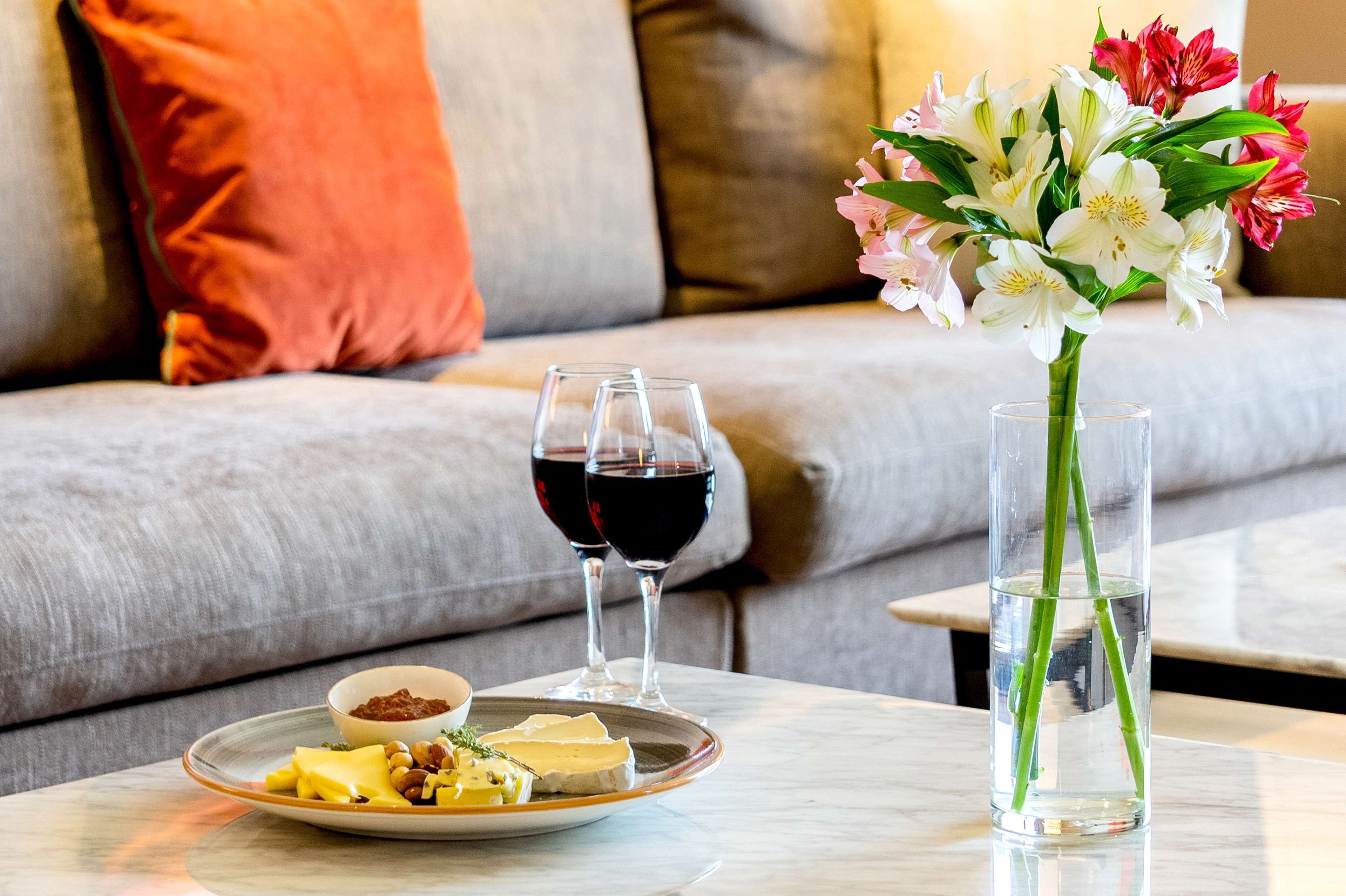 Una mesa de comedor con un plato de queso y frutos secos, dos copas de vino tinto y un florero con flores.