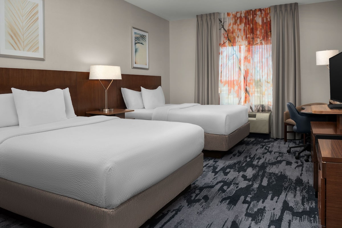 A hotel room with two beds, a desk, a chair, a lamp, and a large window with curtains.