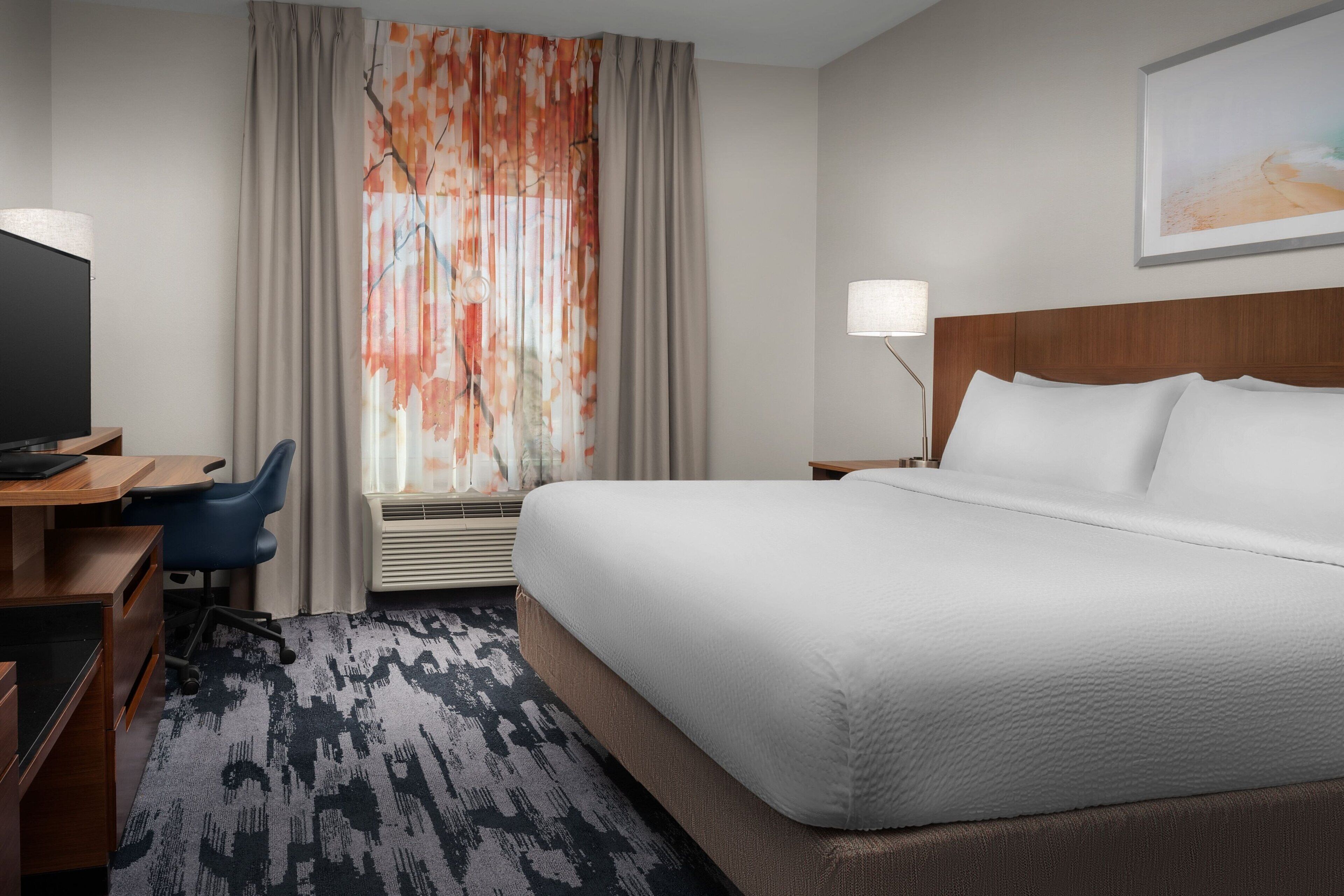 A hotel room with a large bed, a desk with a computer, a chair, and a window with curtains.