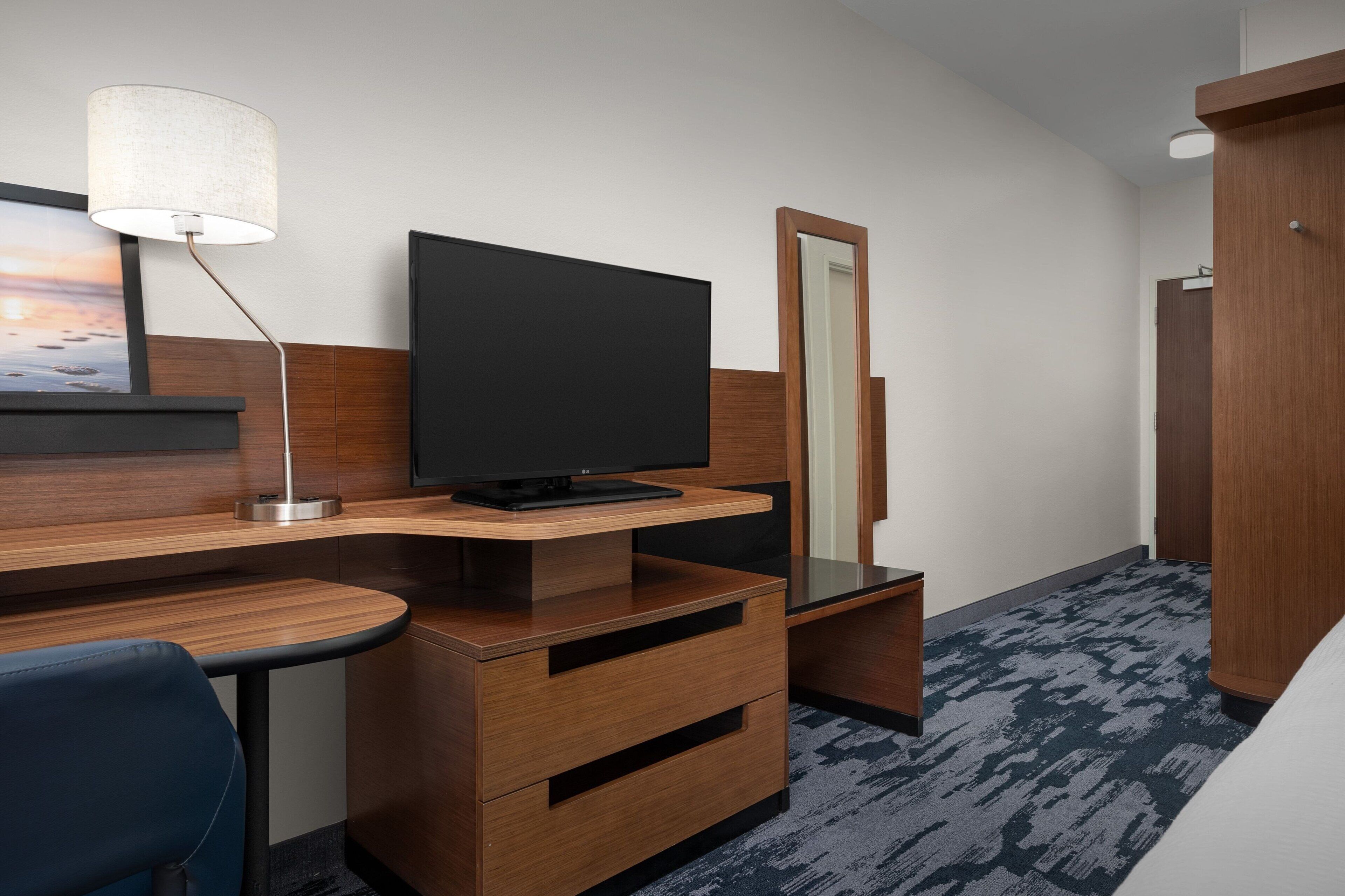 Een hotelkamer met een houten bureau, een flatscreen televisie, een spiegel, een blauwe stoel en een kledingkast.