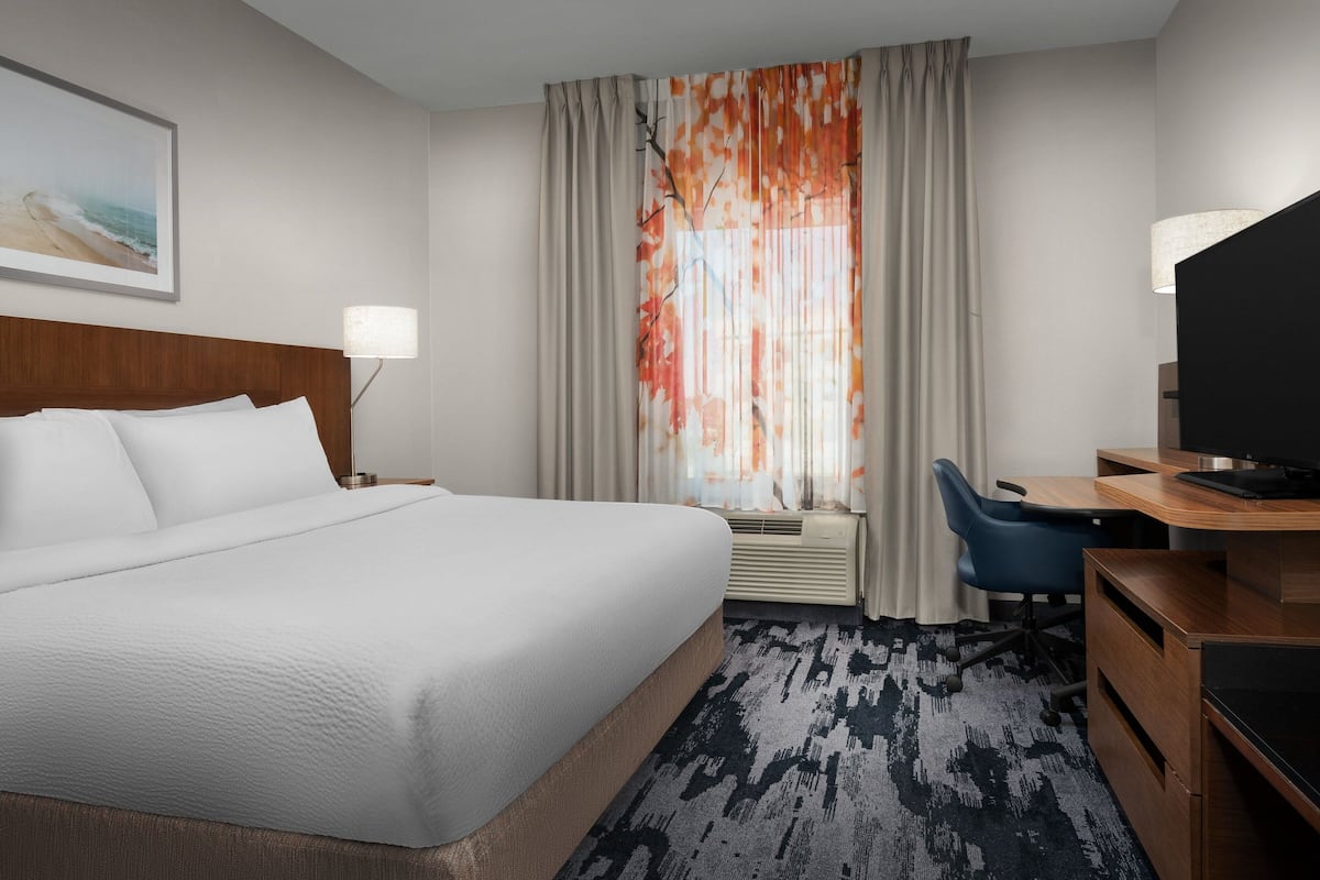 A hotel room with a large bed, a desk with a chair, a television, a window with curtains, and a wall-mounted lamp.