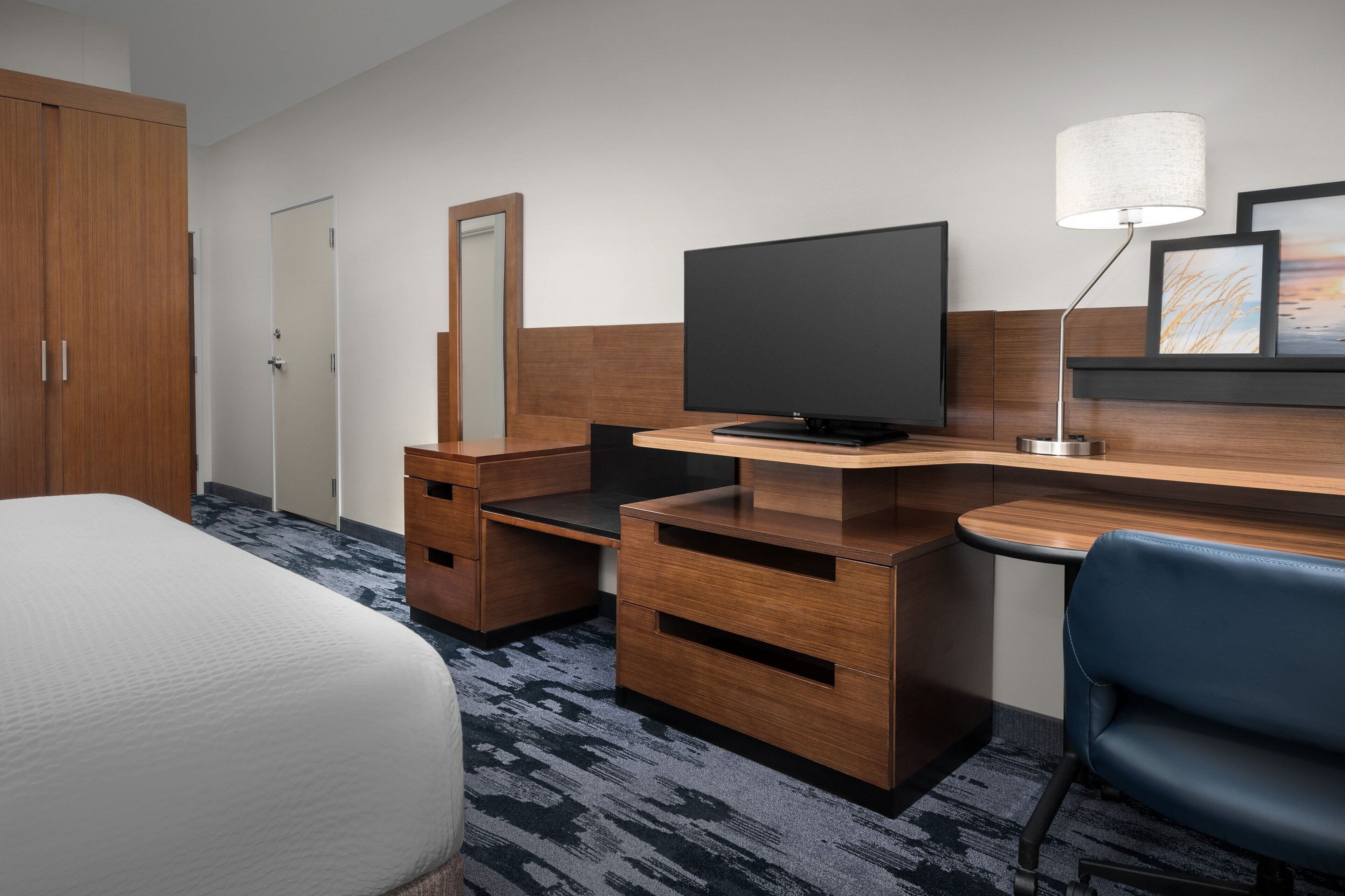 A hotel room with a bed, a desk with a computer, a chair, a TV, and a lamp.