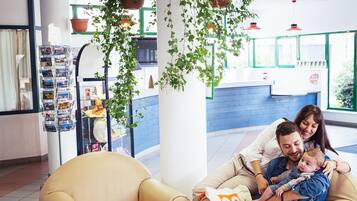 A family sitting on a beige sofa with a pillow labeled "VIVE" in a well-lit indoor space with a reception area in the background.