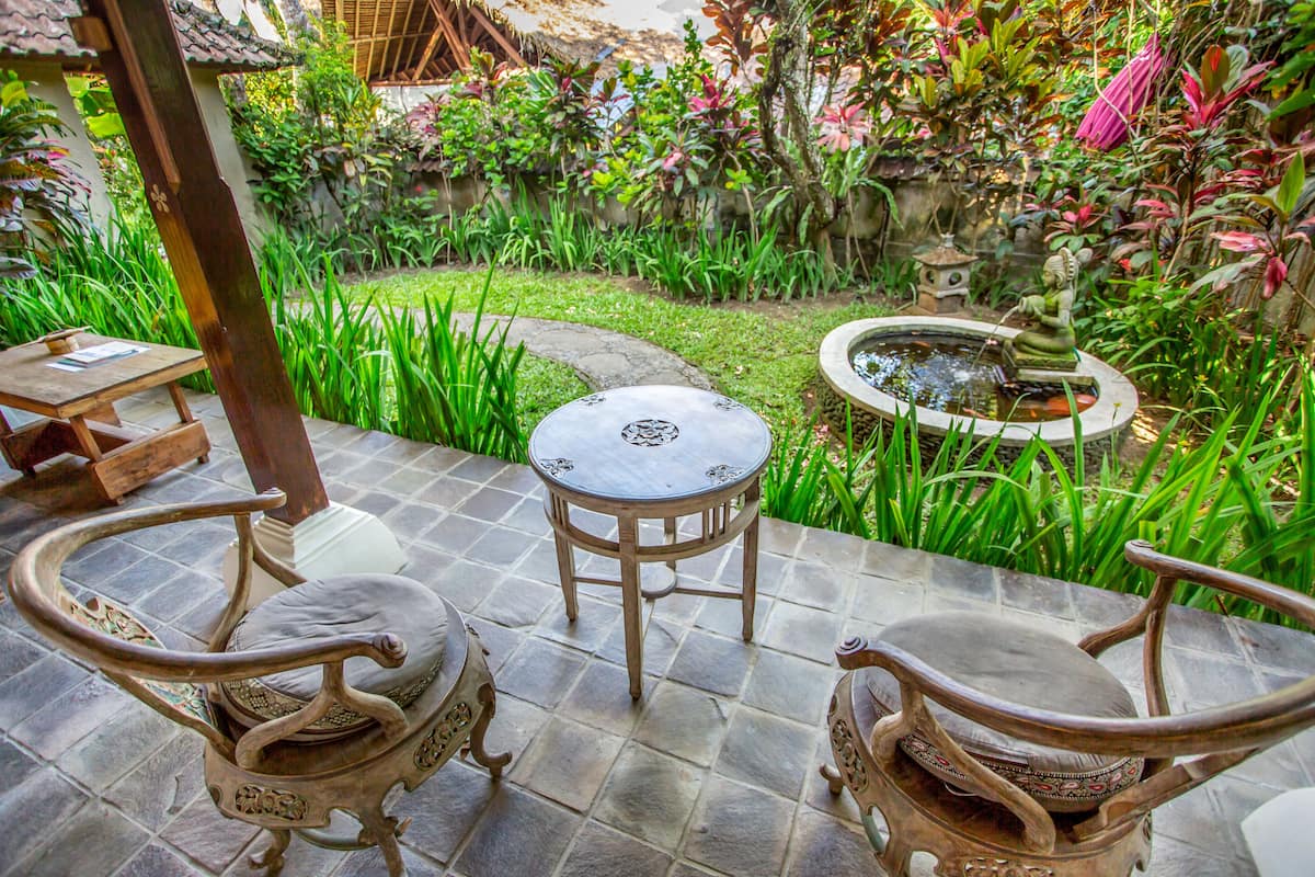 A patio with two ornate chairs and a small table, surrounded by lush greenery and a decorative fountain.