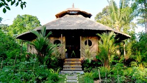 A thatched-roof structure with a central entrance and two circular windows, surrounded by lush greenery. - Swasti Eco Cottages (Ubud)