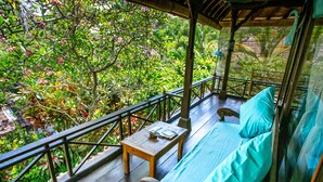 A wooden deck with a sofa and a small table, surrounded by lush greenery and a metal railing. - Swasti Eco Cottages (Ubud)