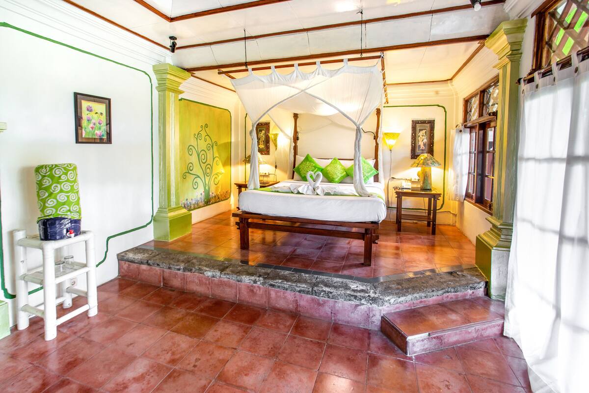 A four-poster bed with a canopy, a small table beside it, and a green patterned chair.